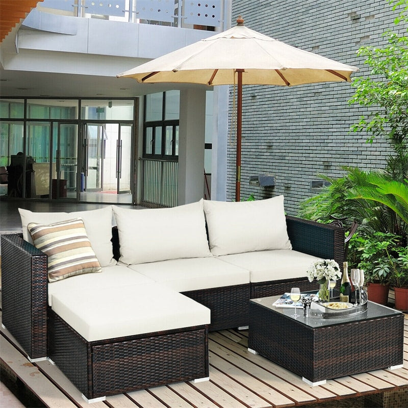 A 5-piece outdoor rattan furniture set with cushions, including a corner sofa, an armless chair, an ottoman, and a coffee table, displayed outdoors with a patio umbrella and plants in the background.